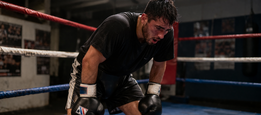 Boxer in ring bent over with hands on knees, breathing heavily after sparring, showing accumulated fatigue