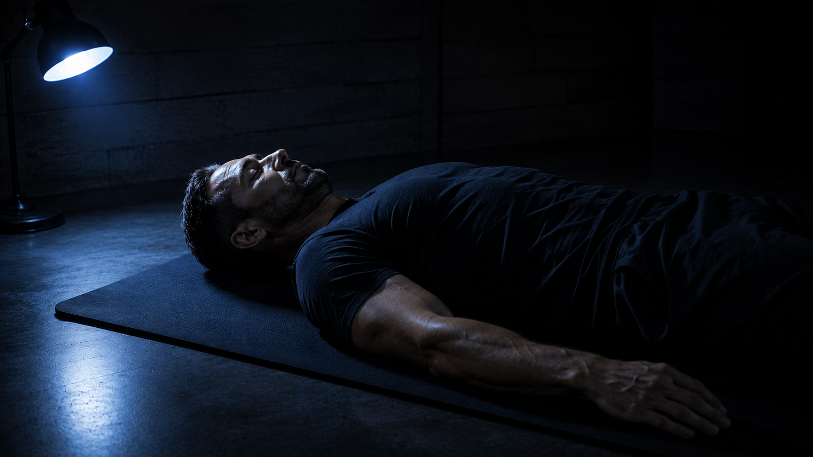 Male athlete in deliberate rest position on training mat, single tungsten lamp, dark quiet room
